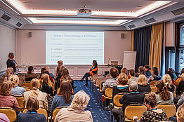 Dipl. Psych. Cäcilia Siemens und Dr. med. Sandra Barth hielten Impulsvorträge im Rahmen des Workshops des Arbeitskreises Psychosoziales Seminarraum mit Zuschauern, vorne sitzen zwei Menschen und simulieren ein Patientengespräch: Dipl. Psych. Cäcilia Siemens und Dr. med. Sandra Barth hielten Impulsvorträge im Rahmen des Workshops des Arbeitskreises Psychosoziales zum Thema: Wie sage ich es? Mit SPIKES? – CF-Behandler als Glücksbringer und Hiobsbotschafter