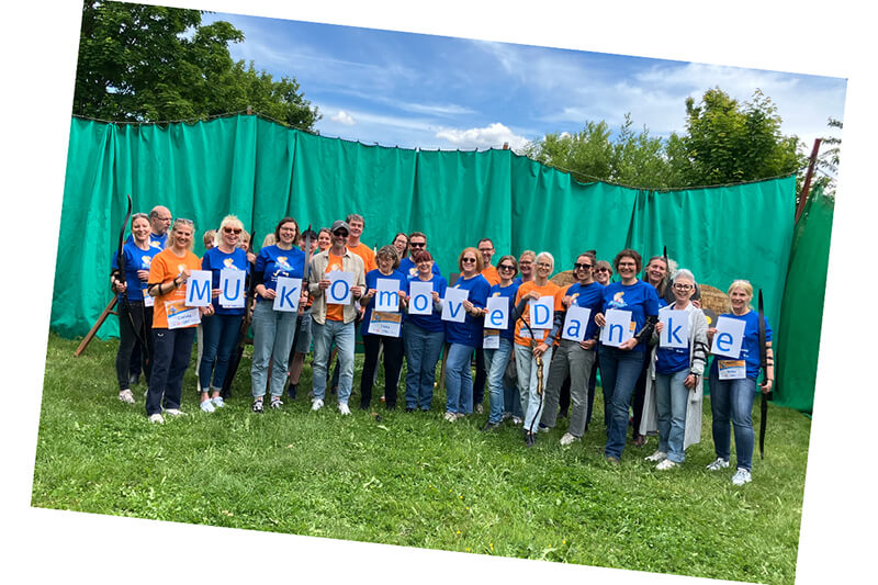 Das Team des Mukoviszidose e.V. sagt Danke für einen tollen MUKOmove 2025. Eine Gruppe von ca. 35 Menschen mit orangenen und blauen MUKOmove-Shirts hält Buchstaben in der Hand, die die Wörter MUKOmove Danke ergeben. Im Hintergrund sieht man eine grüne Plane.